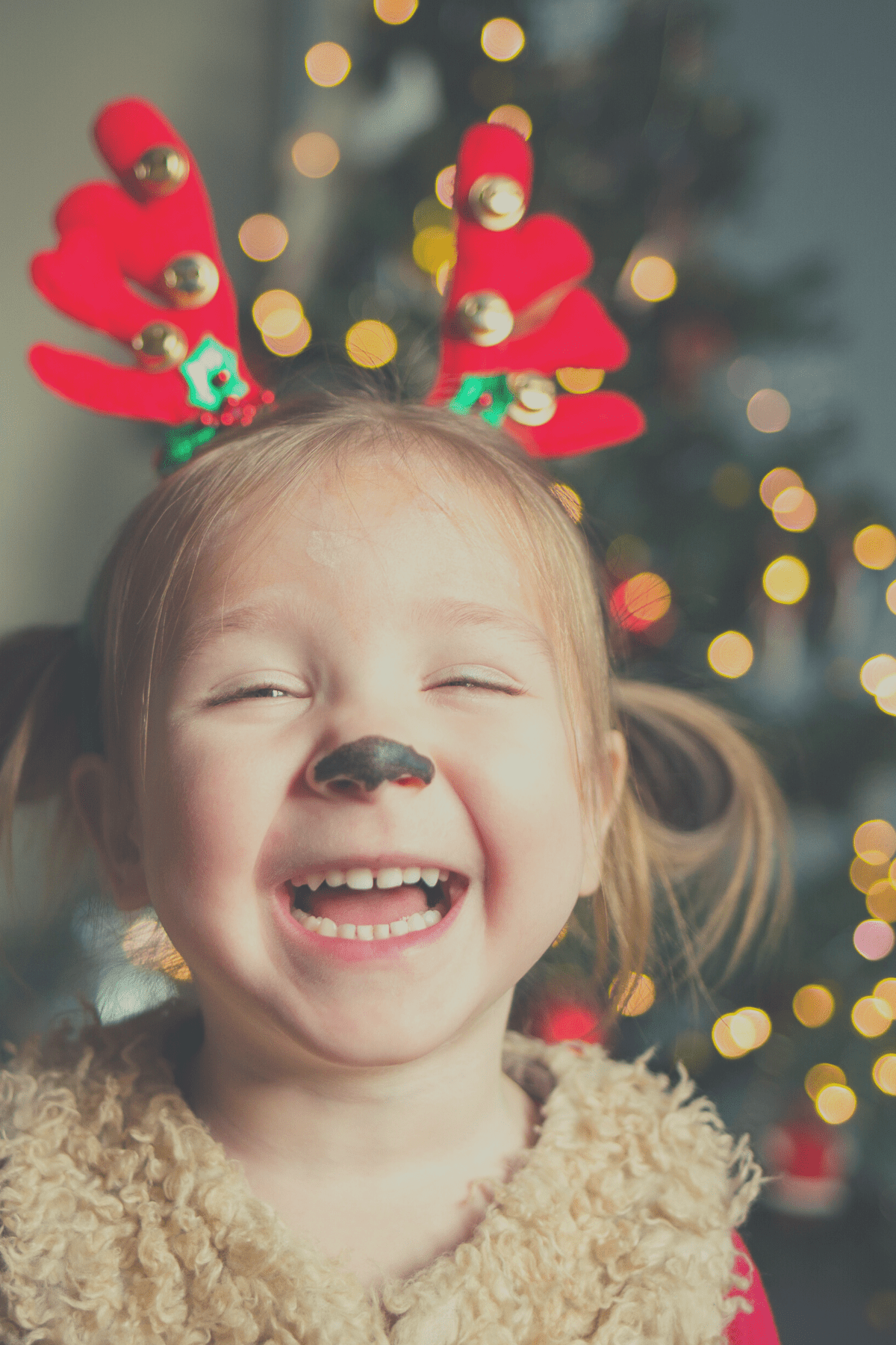 Little girl with holiday antlers and a painted reindeer nose laughing at Would You Rather Christmas questions for kids.