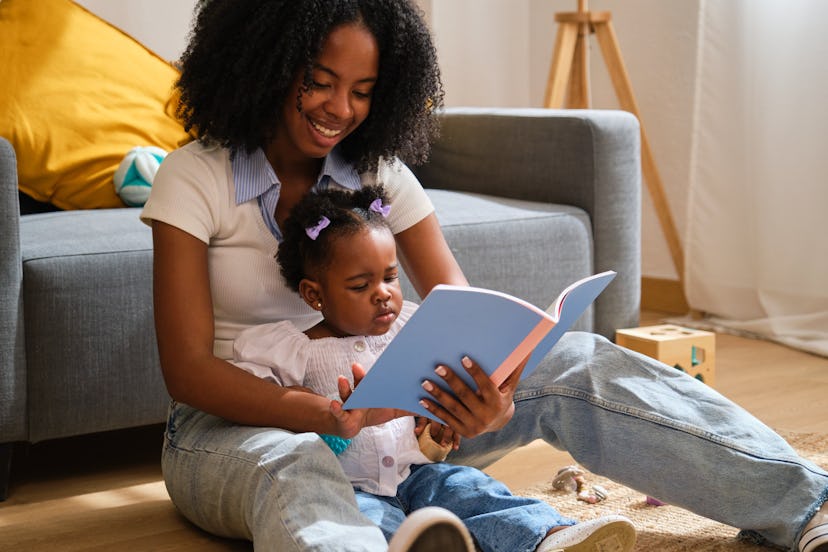 Mom and toddler daughter are sitting on the floor reading a book together