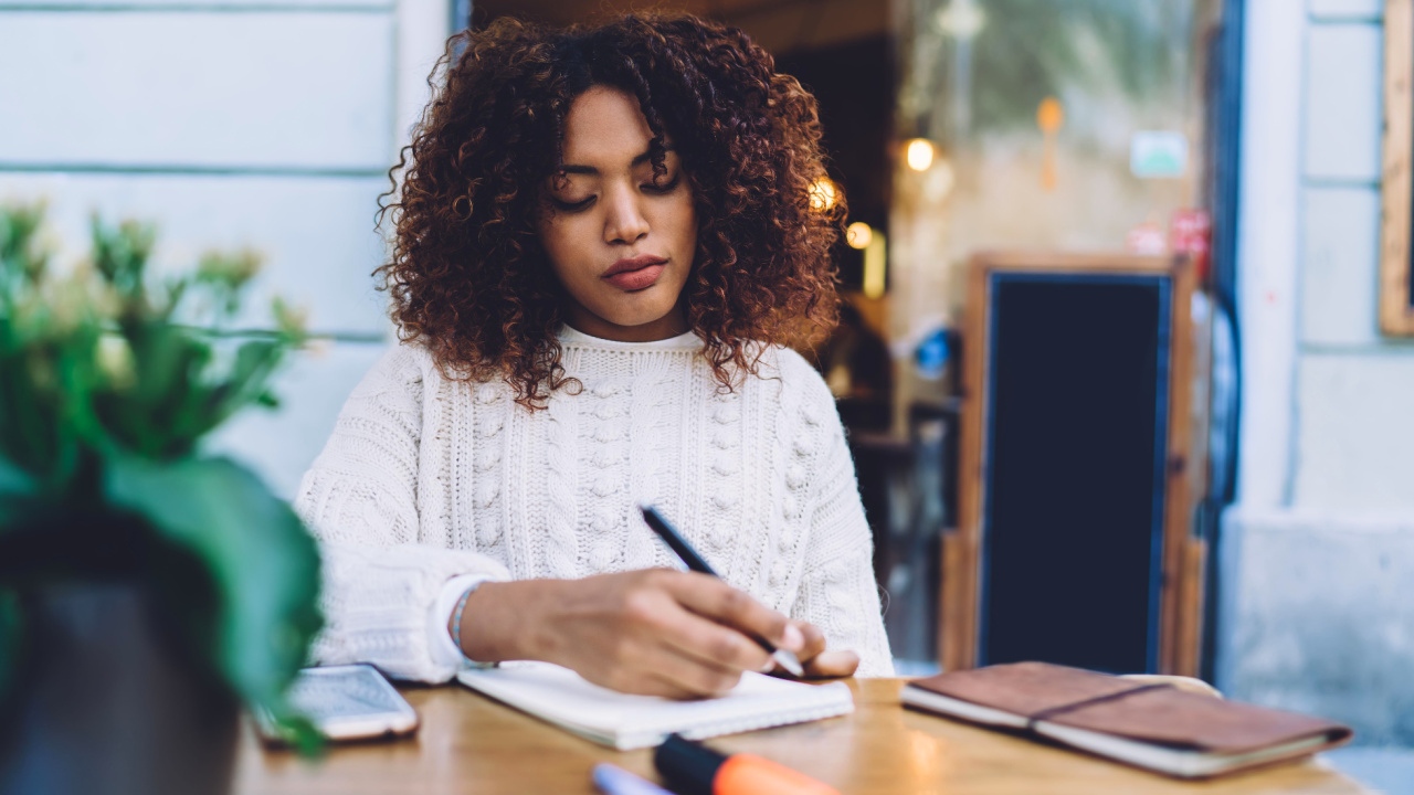 Woman sits at table outside writing in journal.