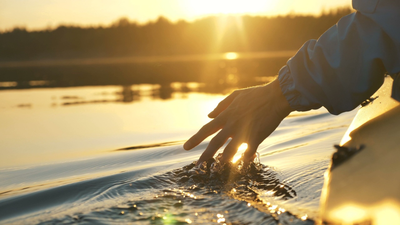 man puts fingers down in lake kayaking against backdrop of golden sunset.