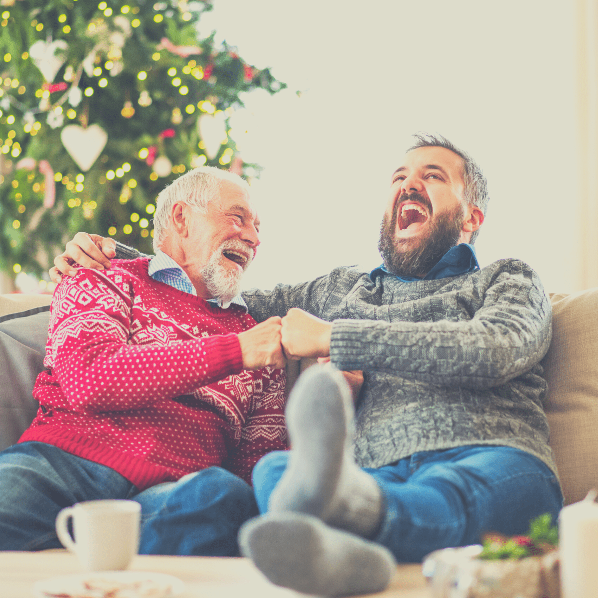 An adult father and son sitting on a sofa laughing and playing Would You Rather Christmas with their family.