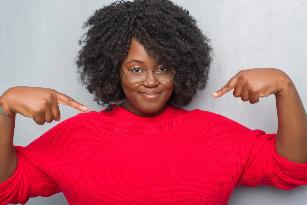 Young african american woman looking confident with smile on face, pointing oneself with fingers proud and happy.