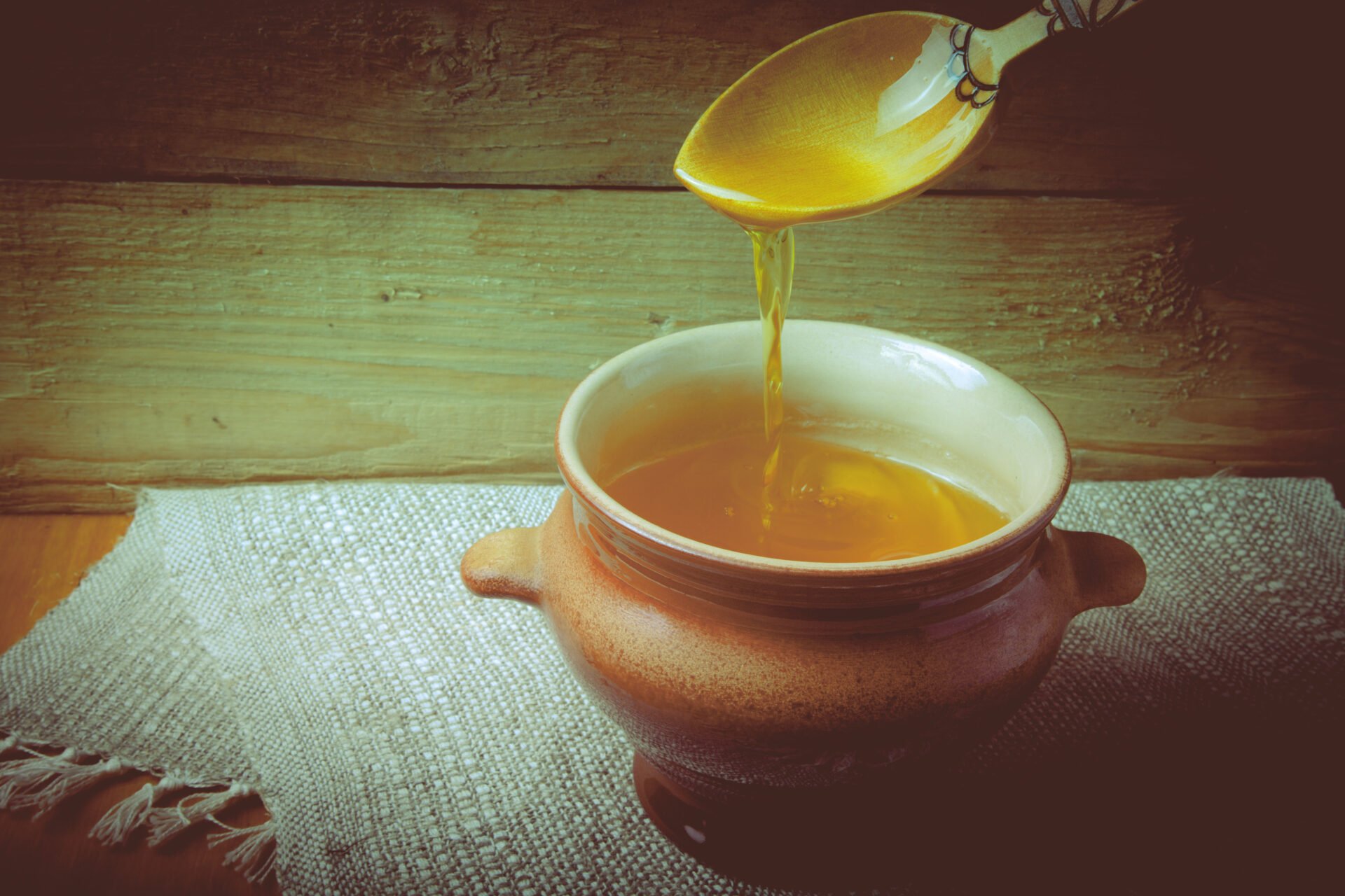 Clay pot with ghee and spoon on linen napkin. Rustic still life. Wooden background.