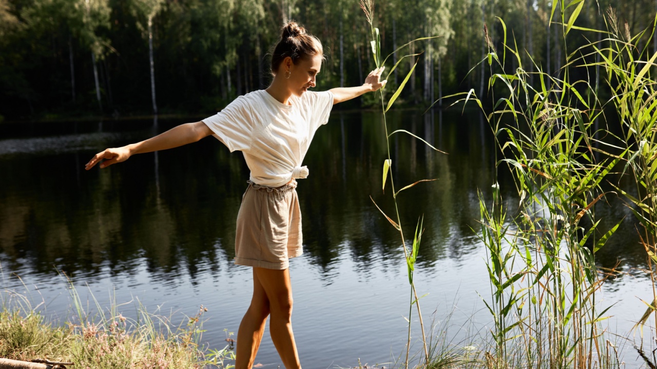 woman outdoors balancing as she walks on a log next to a pond.