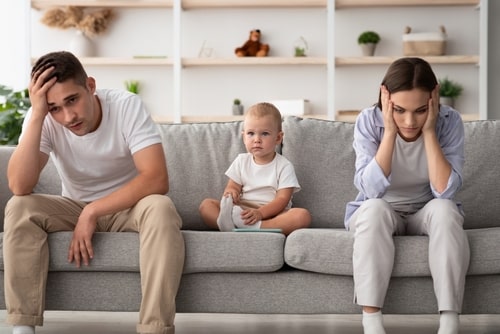 father and mother seated on sofa, each looking exhausted by conflict. Baby sits between them, looking concerned or anxious.