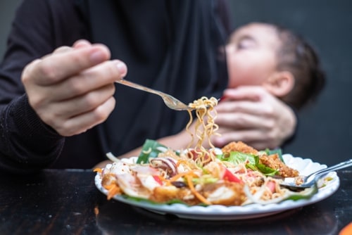 mother's hand holding fork, she's eating from a plate of spicy vegetables and noodles while holding a sleepy infant to her breast